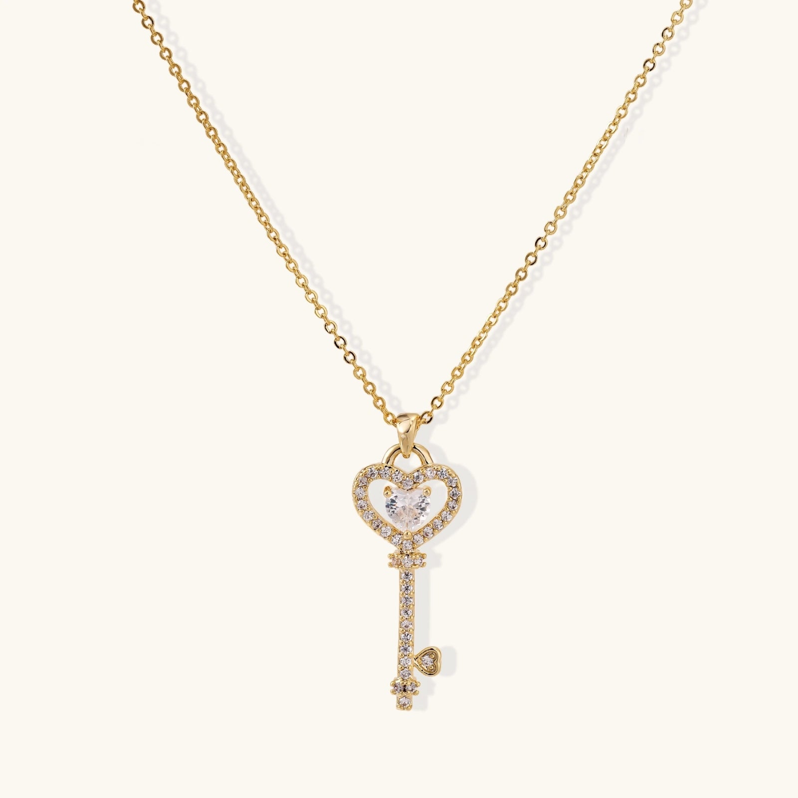 Delicate gold key pendant with heart-shaped bow and crystal pavé detailing on fine chain against cream background.